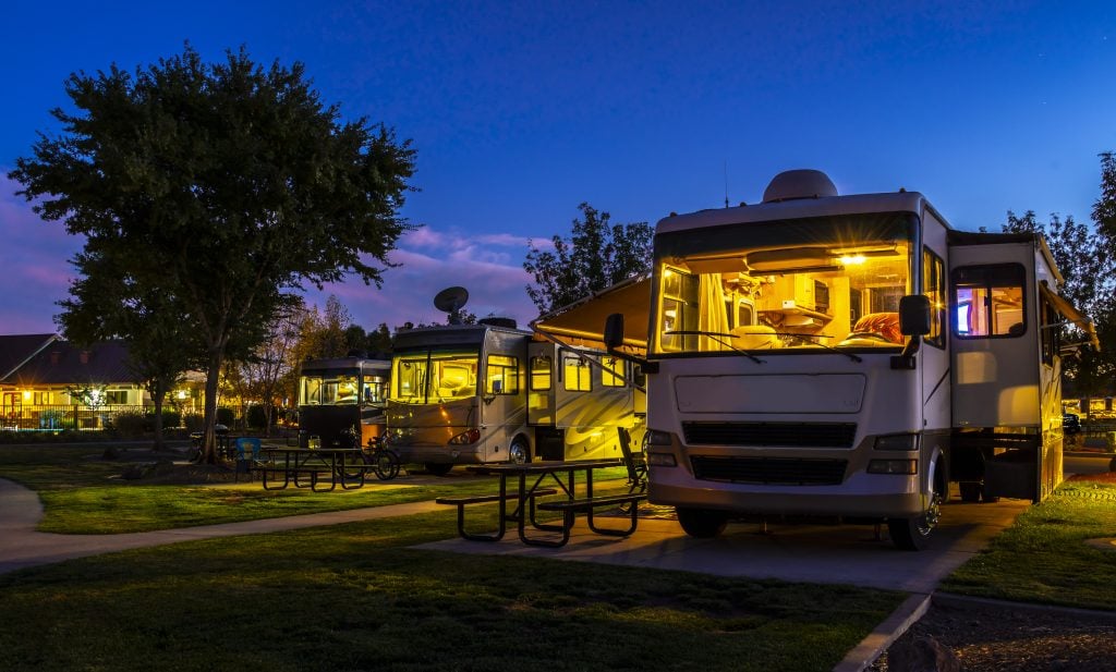 Row of class A RVs in a campground at night with light shining through their big windows. 