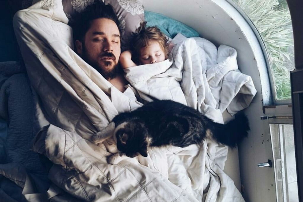 A dad and his small child sleeping in an RV, with the cat sitting on top of both of them. family rv living means having no space. 