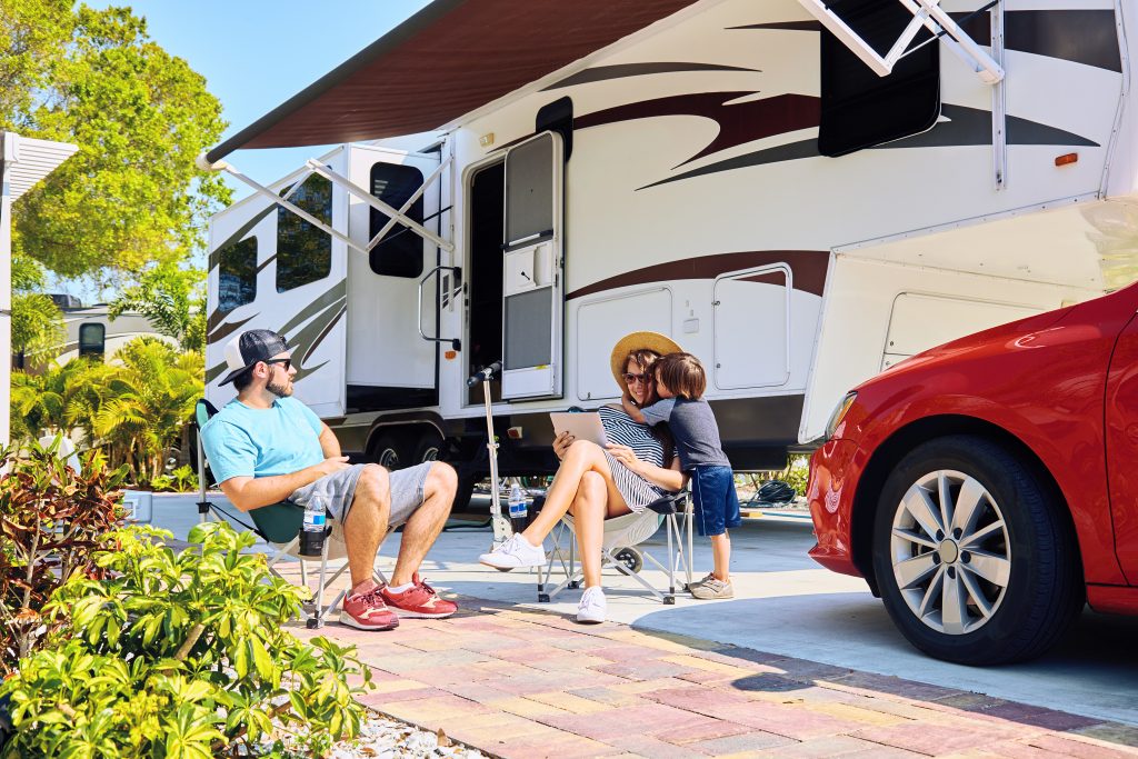 Man and woman sitting in chairs in front of their RV with a child hugging the woman. This family is full-time RV living.
