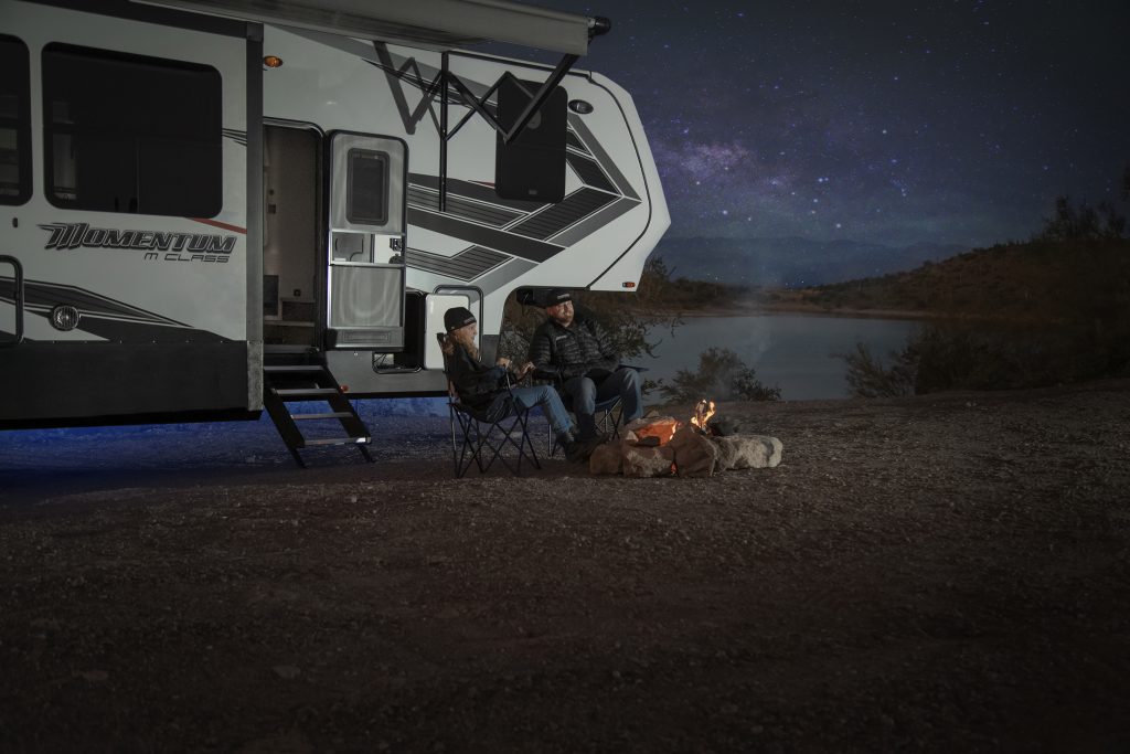 Couple sitting outside of their 5th wheel RV around a campfire at night 