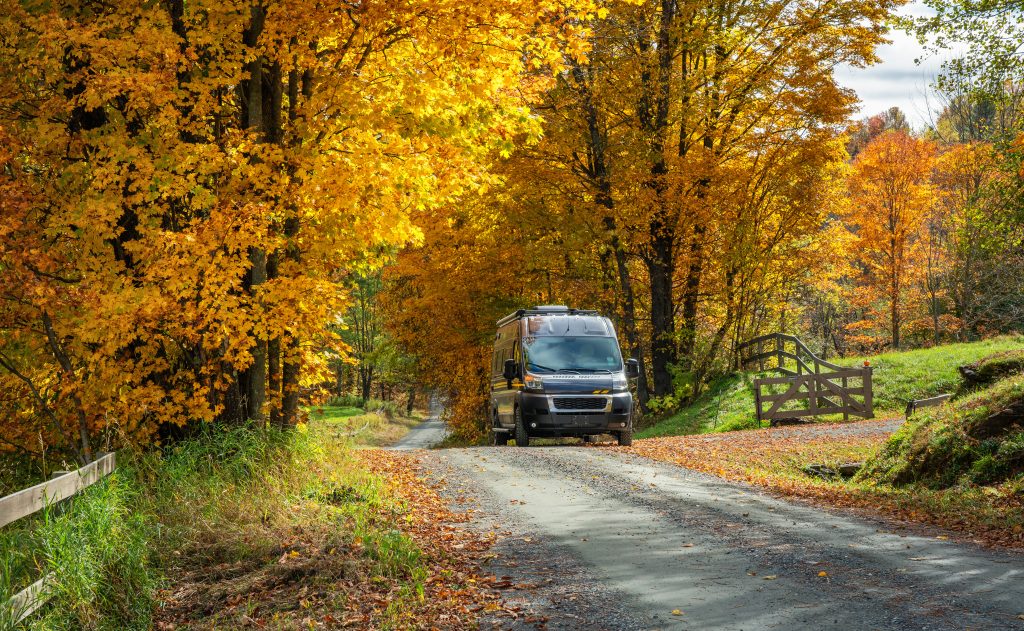 Class B RV driving down a road covered with yellow and orange leaves. 