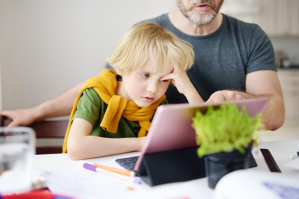 Little boy looking frustrated as his dad helps him with virtual school on his laptop