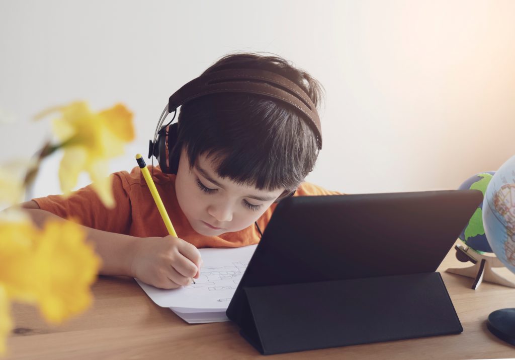 a little boy writing on paper with headphones and his iPad setup on a stand. All items off his virtual school supply list. 