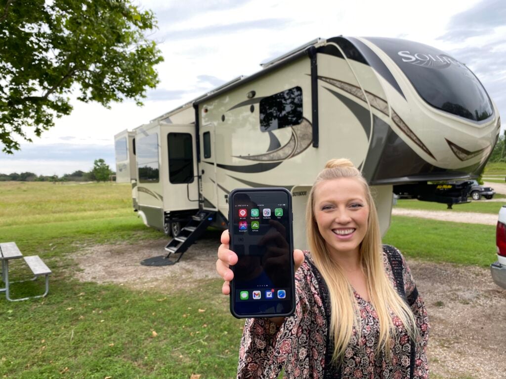 A woman standing in front of her RV, holding her phone showing apps she uses to find RV fresh water fill stations 