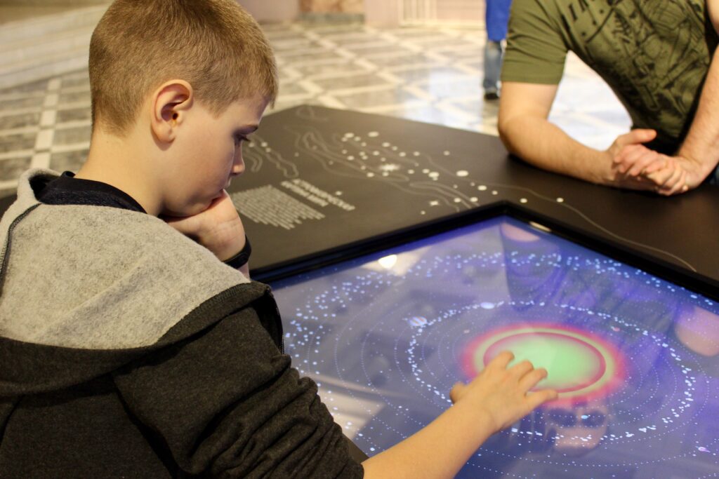 A young boy in a museum playing with a touch screen of planets. Field trips are important to your Roadschooling Schedule