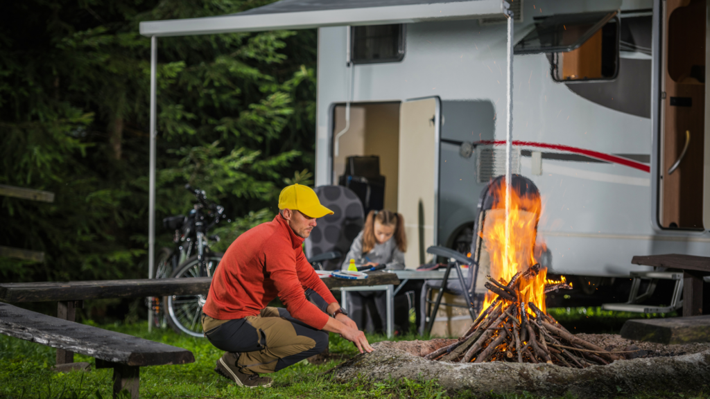 A man looking at the fire with his daughter studying outside of their RV behind him. Part of learning how to travel full time with a family is keeping your kids in school.
