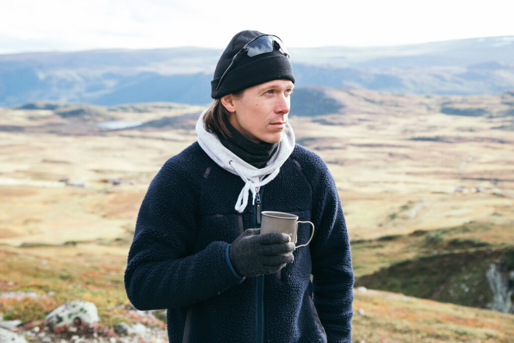 Portrait of young man in warm hiking clothes on epic landscape background, hold titanium metal camping mug, sip coffee at campground.