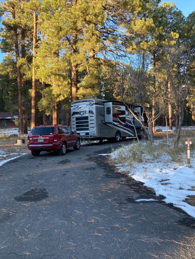 Class A motorhome with red car behind, surrounded by trees with snow on the ground