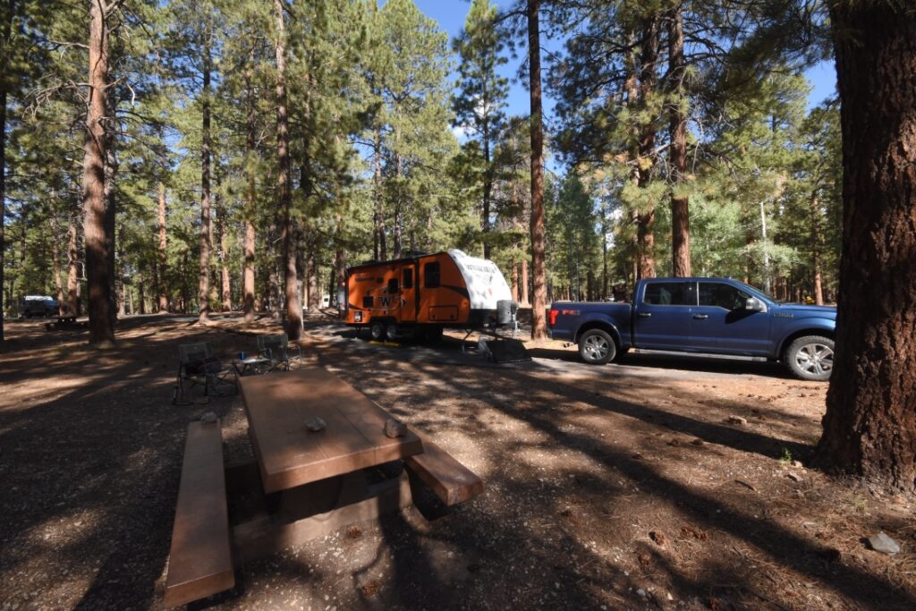 small orange trailer pulled by blue pickup surrounded by woods and picnic table
