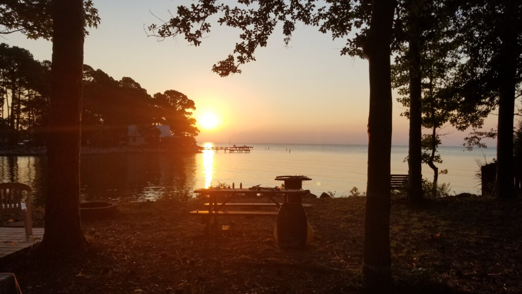View of the sunset over the Rappahannock river. The view is from the back of the wooded campsite with a table and fire pit in the foreground. Feature image for RVing in Virginia at Bethpage and Grey's Point