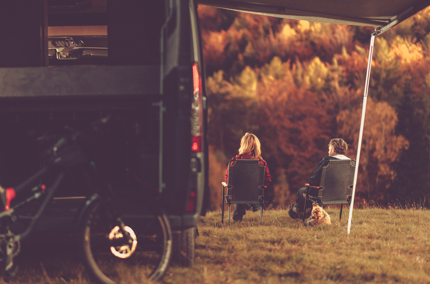 Caucasian Couple and dog in chairs next to their motorhome and Enjoying the fall scenery beside a small rv