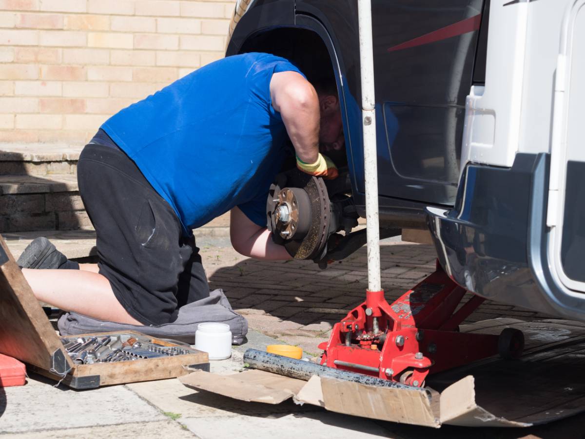 A kneeling mechanic has jacked up a motorhome recreational vehicle and removed a wheel and peers under the wheel arch checking the brakes.Trolley jack, socket set are visible