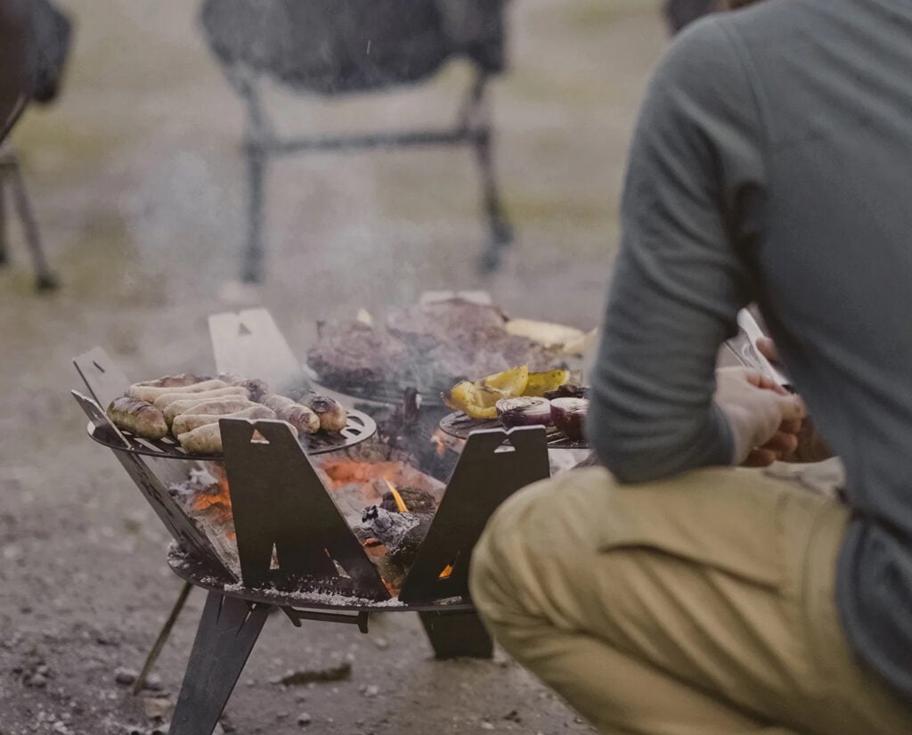 Man cooks brats and vegetables on Firebrand RV fire pit and grill.