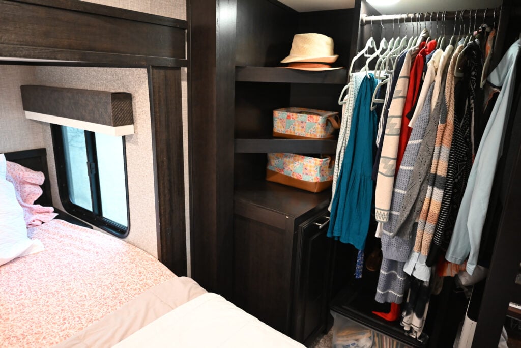 a closet filled with lots of clothes next to a bed and a hat on top of a wooden shelf, packing