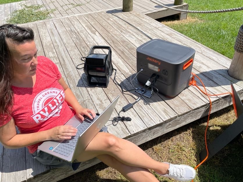 Woman sits on deck with laptop and portable fan, both plugged into the Jackery 2000 Plus