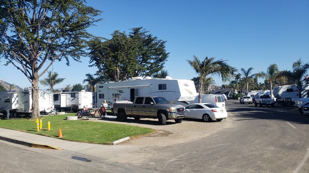 Pismo Coast Village RV Park campsite with grassy area and trees. A fifth-wheel and two cars are parked next to it. Palm trees in the background.