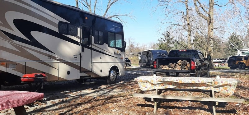 white and black Class A camper parked next to a black pickup truck with firewood in the bed and picnic table in the foreground