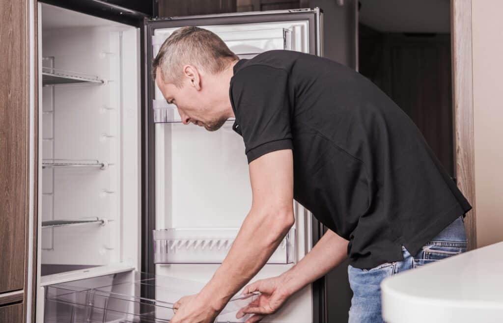 A man with an RV fridge door open is holding one of the shelves while cleaning the interior.